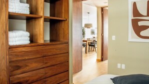 Een houten garderobe met opgevouwen handdoeken, een bed met een kussen, en een eethoek op de achtergrond.