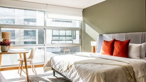 Ein modernes Hotelzimmer mit einem großen Bett, einem Schreibtisch und Blick auf die Stadt durch bodentiefe Fenster.