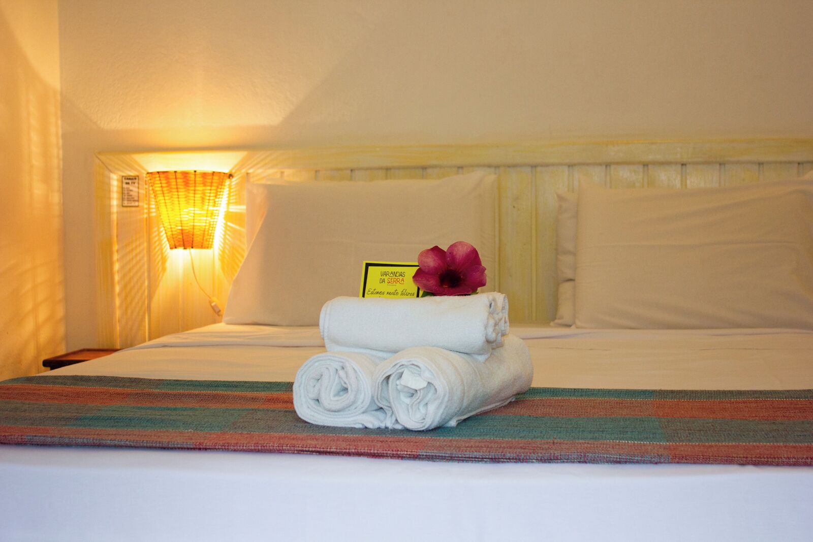 A neatly made bed with rolled towels and a flower, a wall-mounted lamp, and a sign with text.