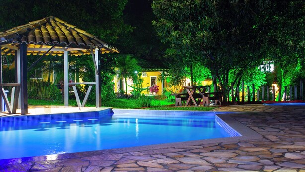 Un área de piscina nocturna con un deck empinado, un pabellón y un jardín bien iluminado con árboles y una mesa.