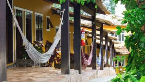 Un espacio al aire libre techado con hamacas colgando de pilares de madera, suelo de piedra y un edificio amarillo con ventanas y contraventanas.