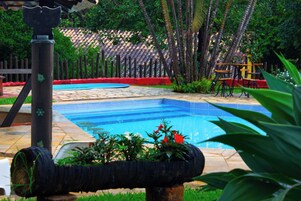A swimming pool with a tiled deck, surrounded by greenery and a wooden fence.