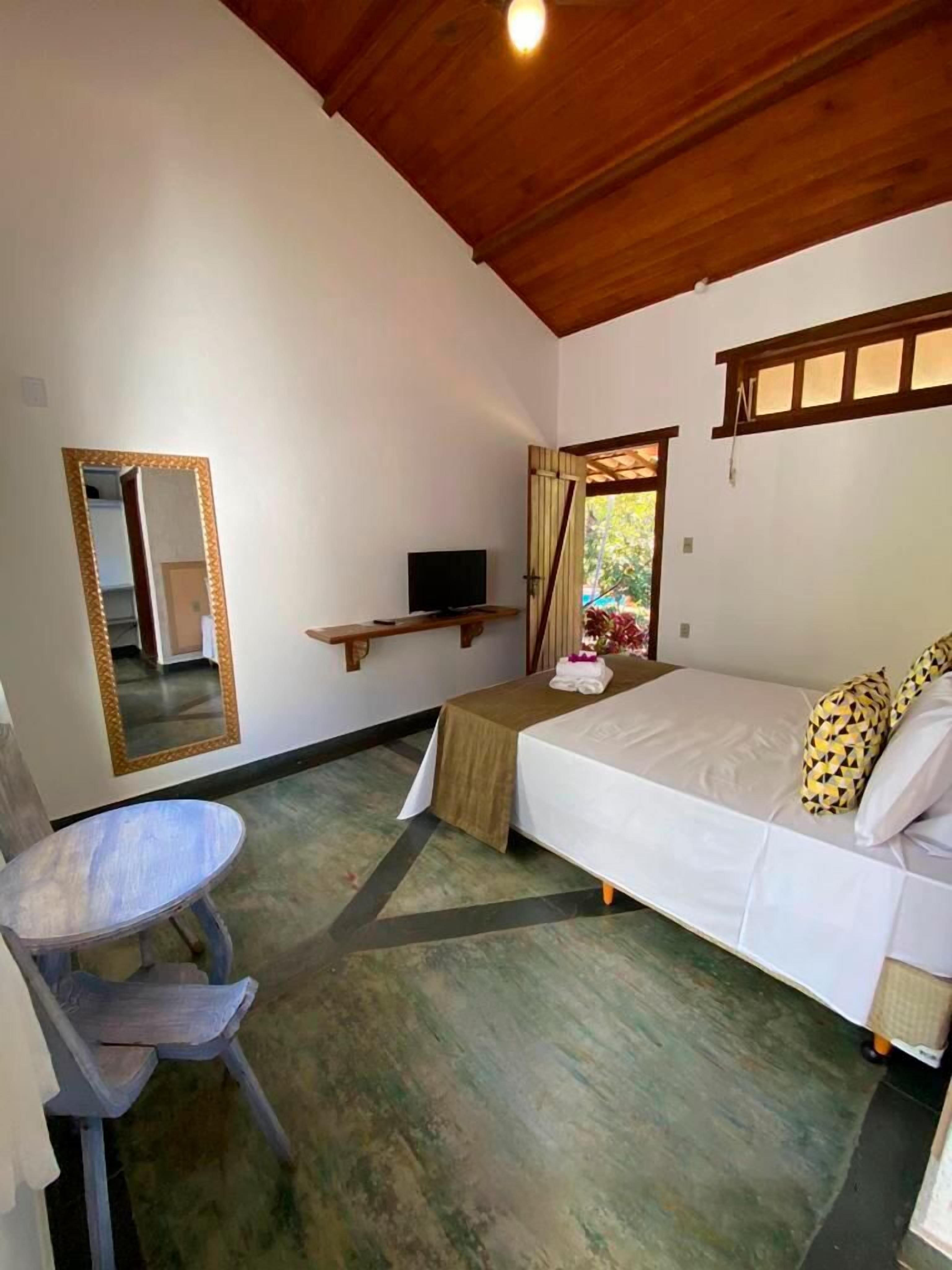 Quarto com uma cama, uma mesinha, uma cadeira, uma televisão e um espelho.