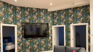 A living room with a large flat-screen TV mounted on the wall, a grey sofa with colorful cushions, a small white table with a bowl and spatula, and a wooden floor. - Apartment 5 (York)