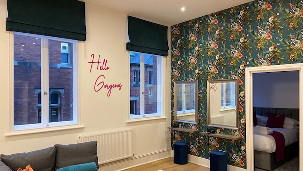 A modern living room with a grey sofa, a coffee table, and a bed with red pillows. A wall with a floral wallpaper and a 'Hello Gorgeous' sign is visible. - Apartment 5 (York)