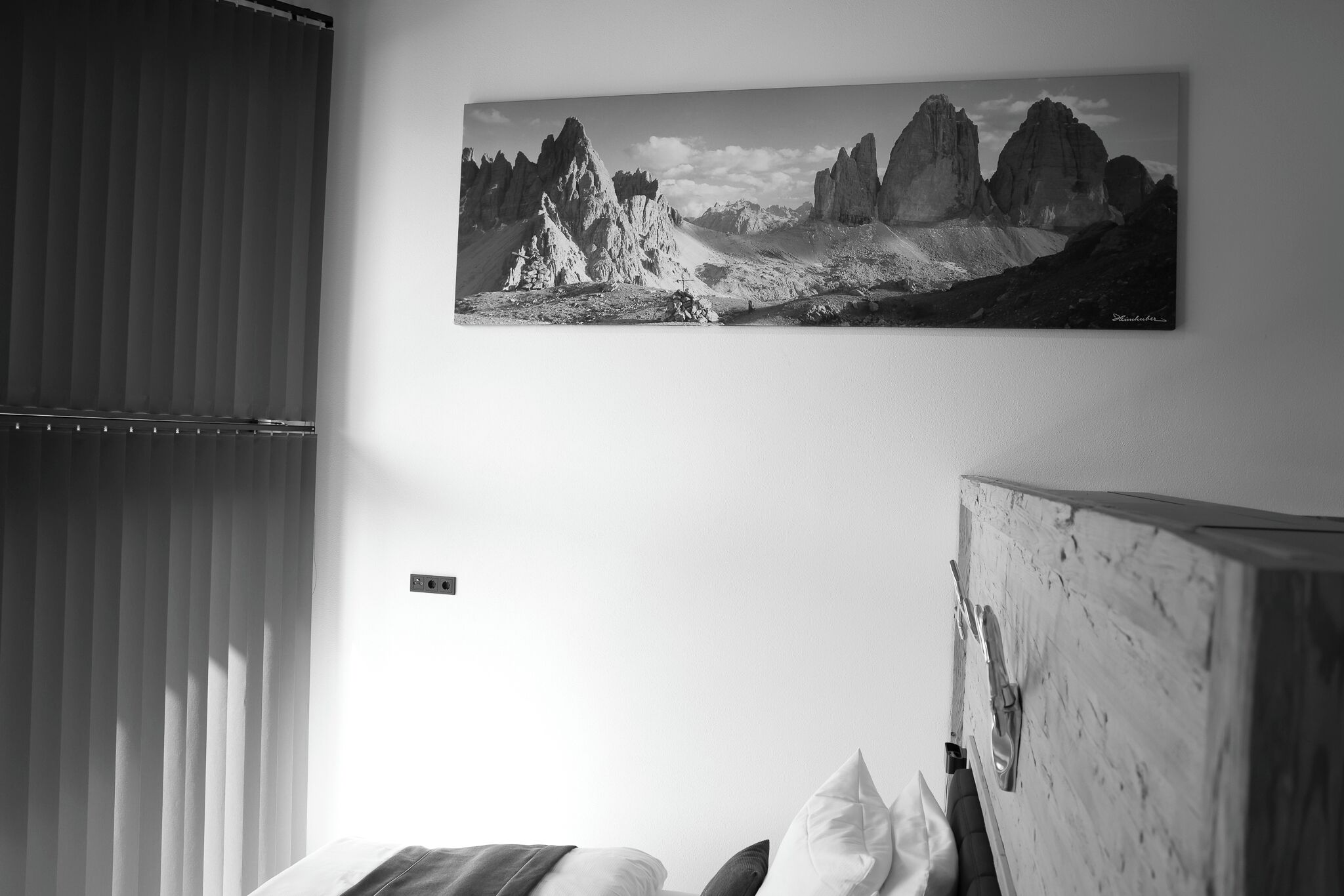 A bedroom with a bed, a window with blinds, and a mountain landscape painting on the wall.