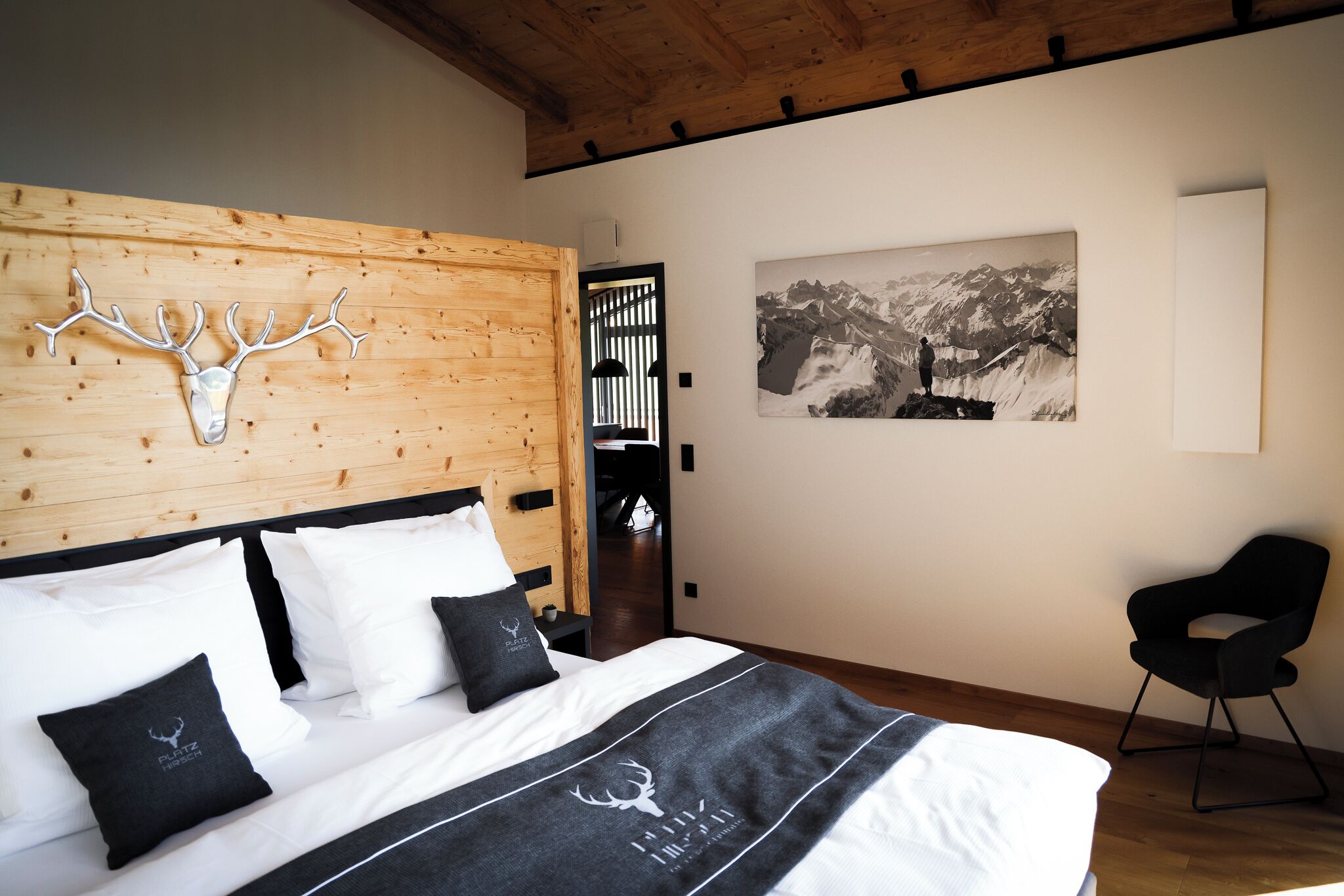 Ein Schlafzimmer mit einem hölzernen Kopfteil, einem Bett mit weißer und schwarzer Bettwäsche, einem aufgehängten Hirschkopf, einem schwarzen Stuhl und einem Gemälde einer Berglandschaft an der Wand.