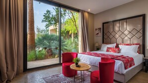 Ein modernes Hotelzimmer mit einer großen Schiebeglaswand zum Garten, einem Bett mit rot-weißer Bettwäsche und roten Stühlen.