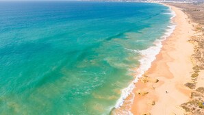 Vista aérea de una playa con aguas turquesas cristalinas, orilla arenosa y edificios a lo lejos.