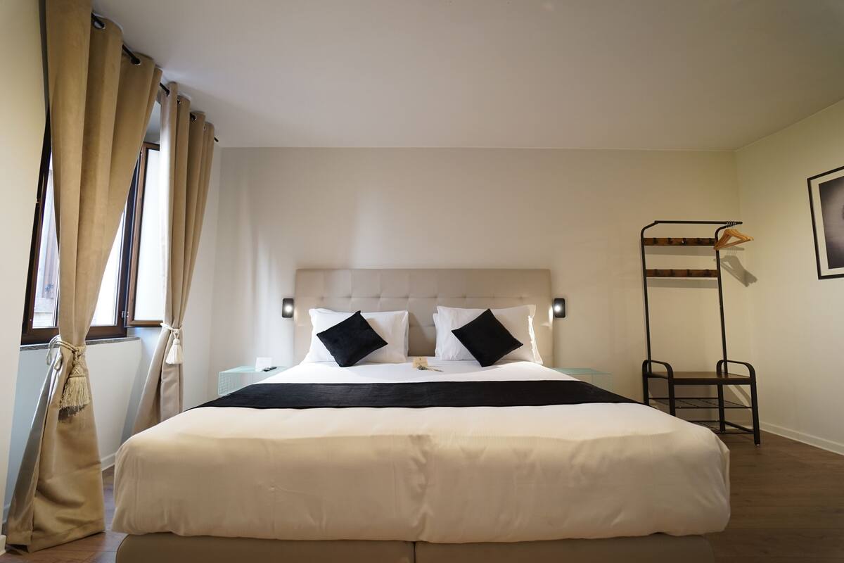 Ein ordentlich eingerichtetes Hotelzimmer mit einem großen Bett, einem Nachttisch, einem Hängesessel und einem Fenster mit Vorhängen.