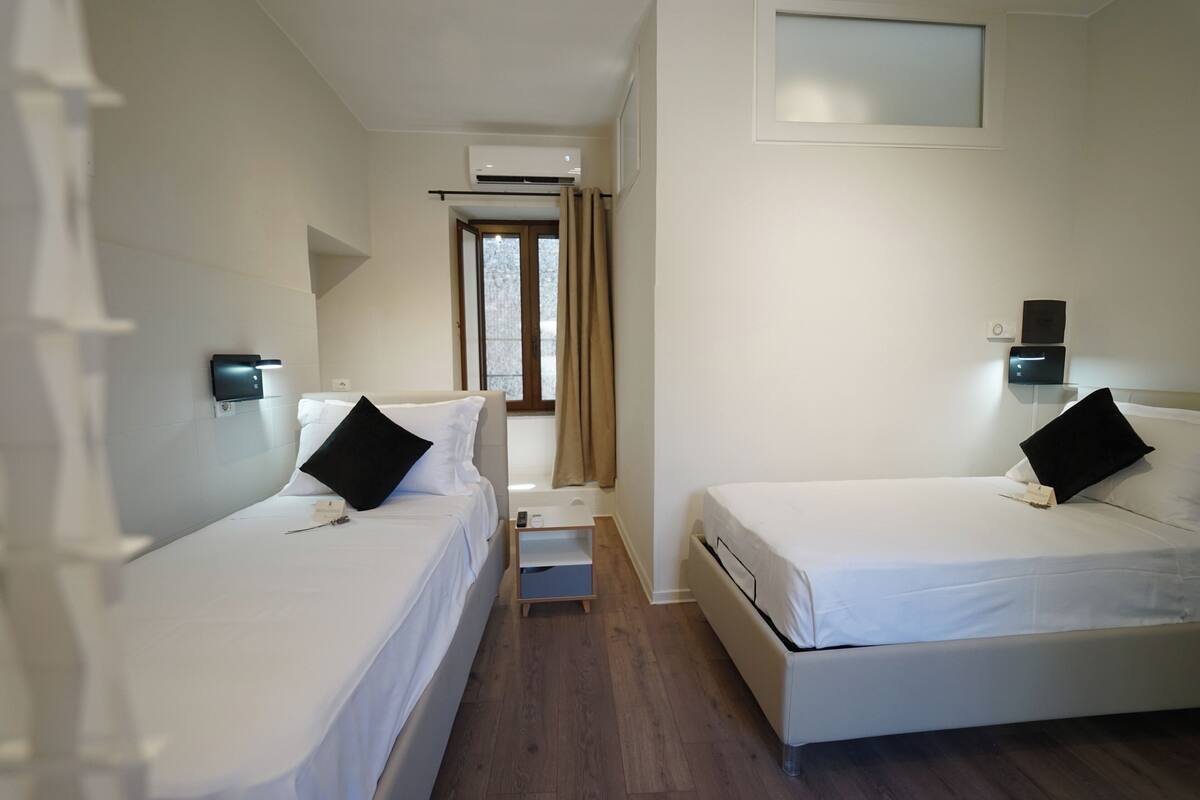 Ein Hotelzimmer mit zwei Einzelbetten, einem kleinen Nachttisch, einer Wand-Klimaanlage und einem Fenster mit Vorhängen.