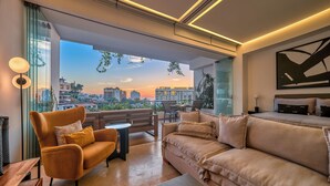 Een moderne woonkamer met een bank, fauteuil en salontafel. Er is een groot raam met uitzicht op gebouwen en een zonsondergang.