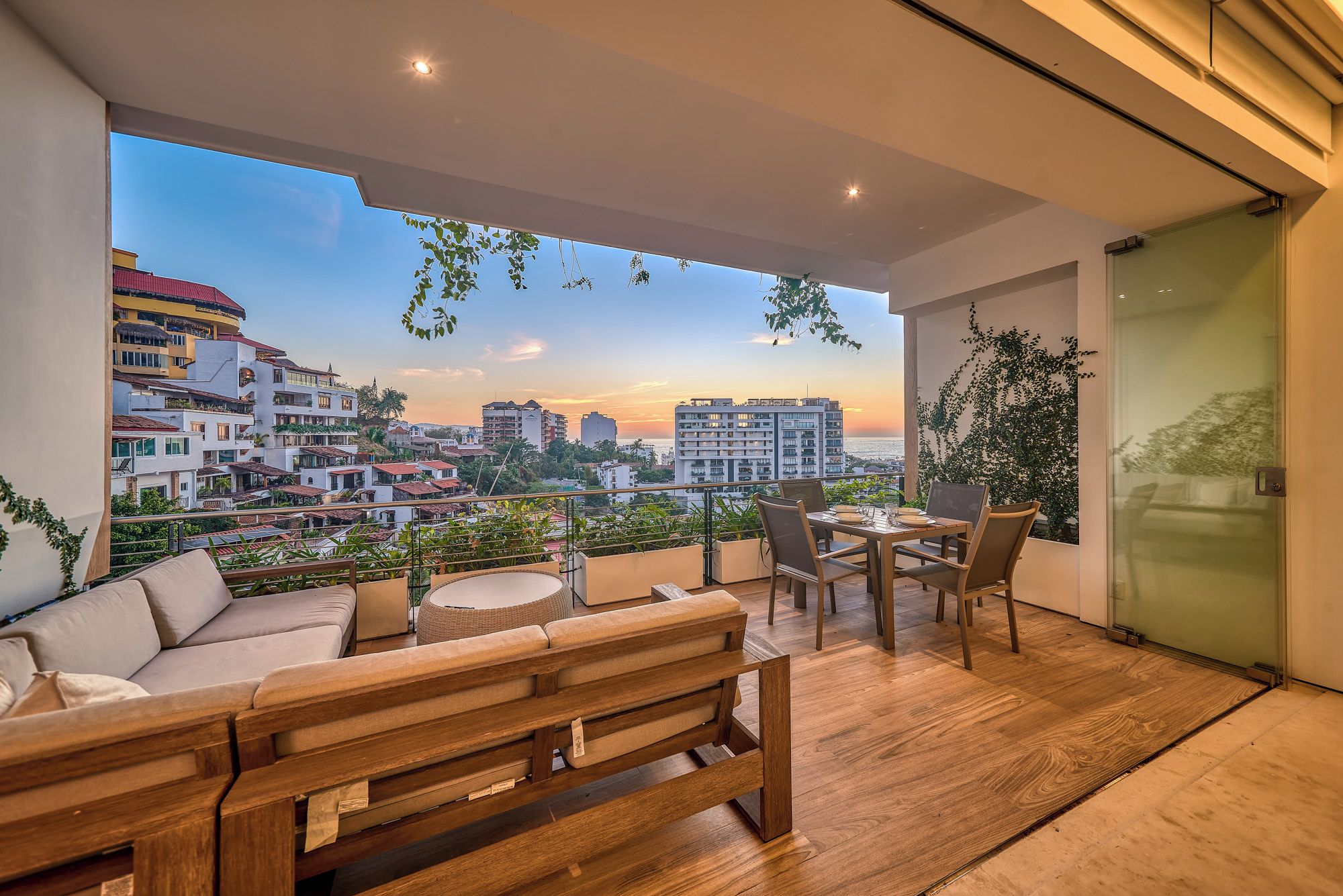 Ein Balkon mit einer Holzbank, einem Tisch und Stühlen mit Blick auf eine Stadtlandschaft bei Sonnenuntergang.