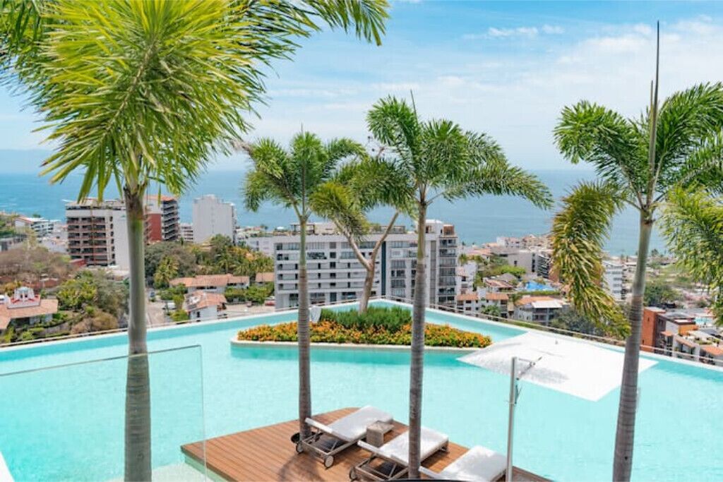 Zona de piscina con palmeras, sillas de descanso y vistas a la ciudad.