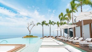 Ein Poolbereich mit Liegestühlen und Sonnenschirmen, Palmen und Blick auf den Ozean.