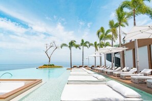 Un espace aménagé au bord de la piscine, avec des chaises longues et des parasols, des palmiers et une vue sur l’océan.