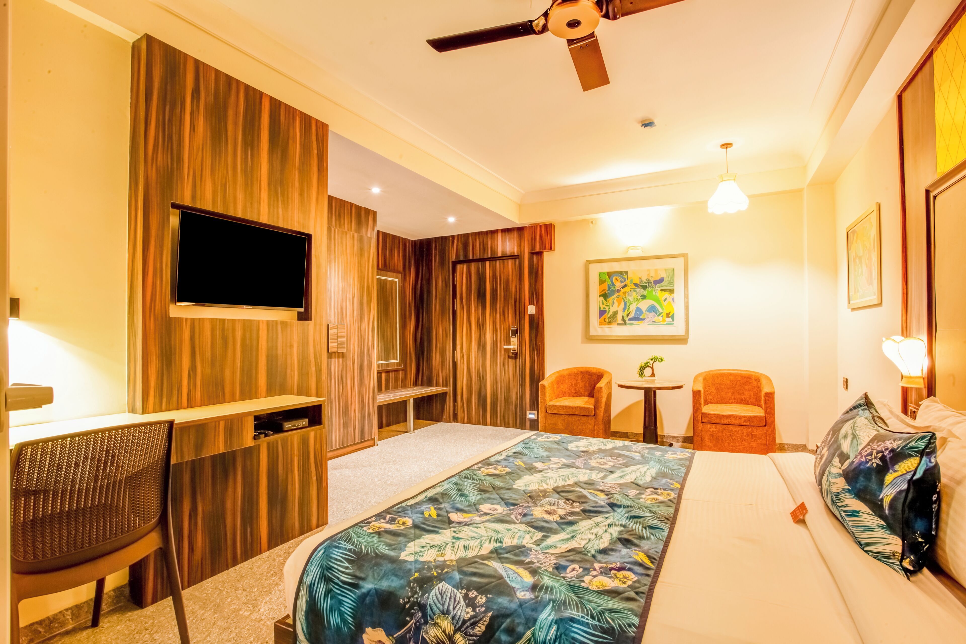 Una habitación de hotel con cama, dos sillas, escritorio, televisor y ventilador de techo.