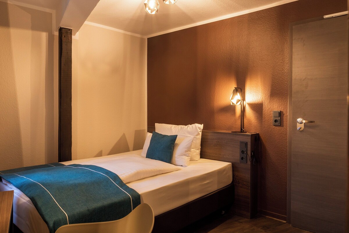 Una habitación de hotel con una cama individual, una mesita de noche, una lámpara y una puerta.