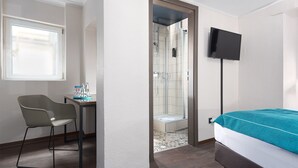 Habitación de hotel con cama, escritorio con silla, zona de ducha y televisor montado en la pared.