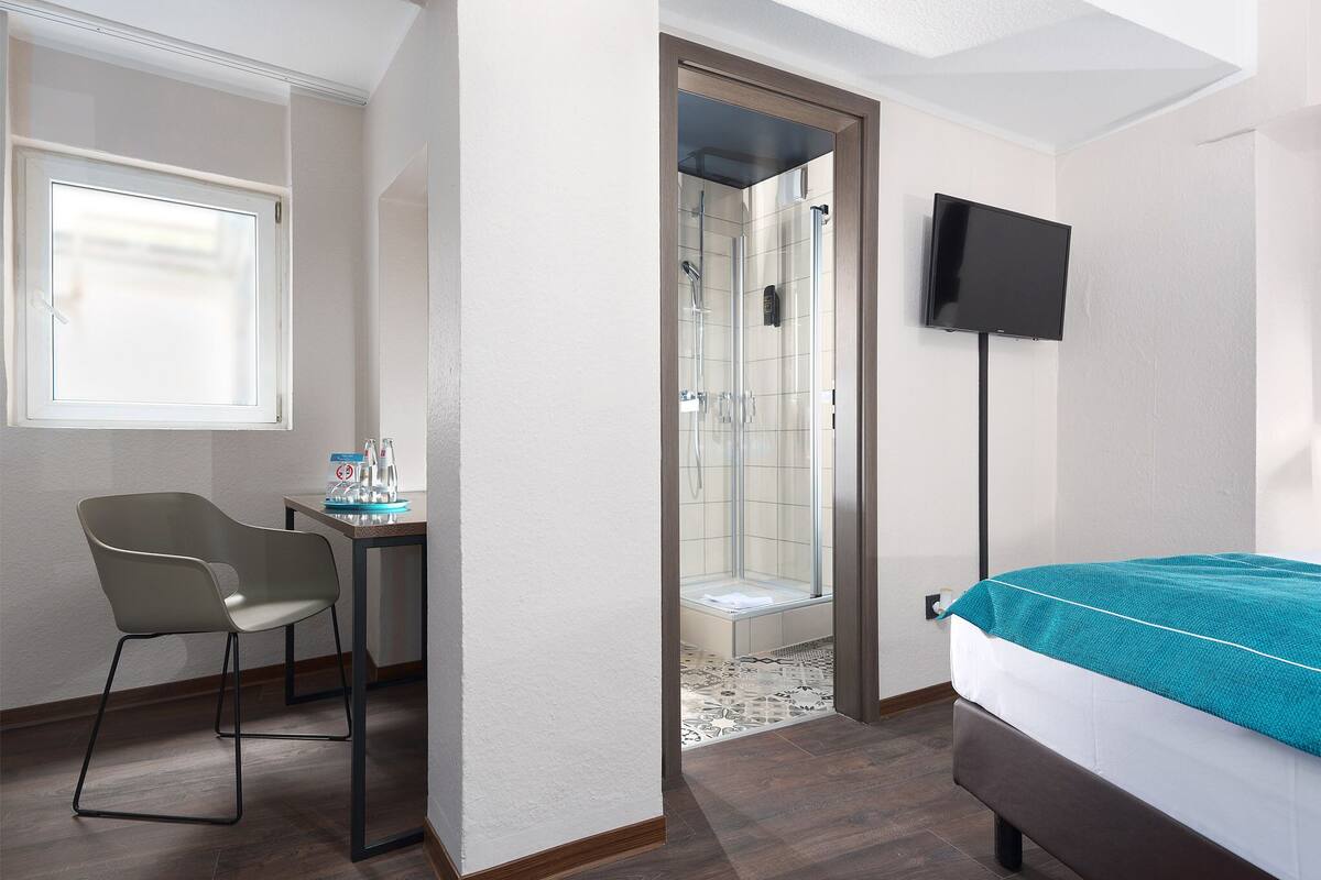 A hotel room with a bed, a desk with a chair, a shower area, and a TV mounted on the wall.