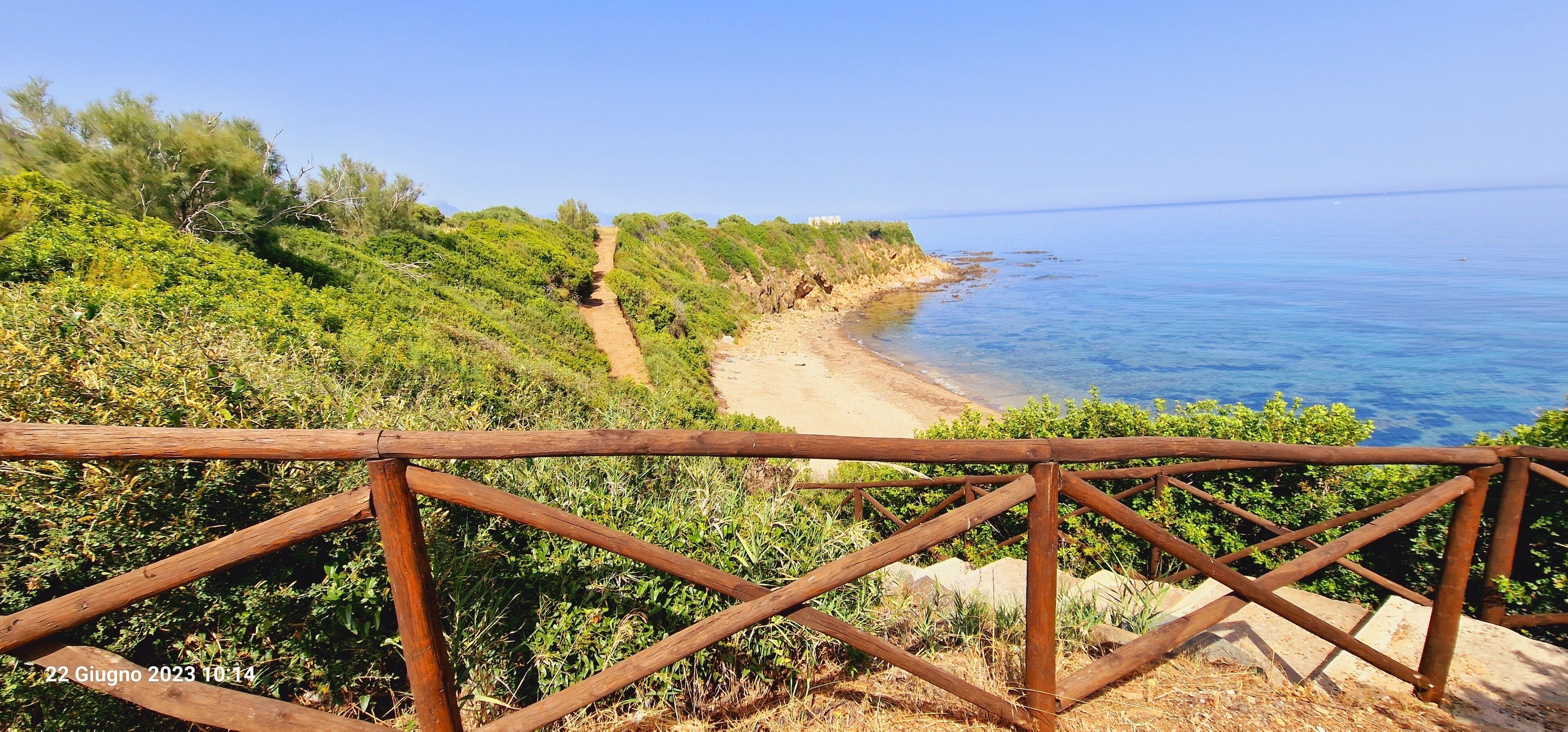 Paisaje costero con un sendero de tierra que lleva a una playa, rodeado de vegetación y una cerca de madera.
