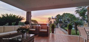 A covered outdoor seating area with wicker furniture, a potted plant, and a view of the sunset.