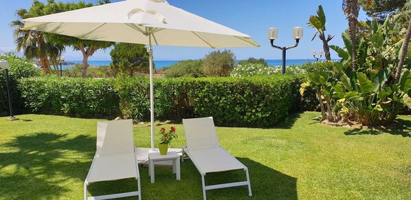 A garden with two white lounge chairs and a table under a white umbrella, surrounded by greenery and a view of the sea.