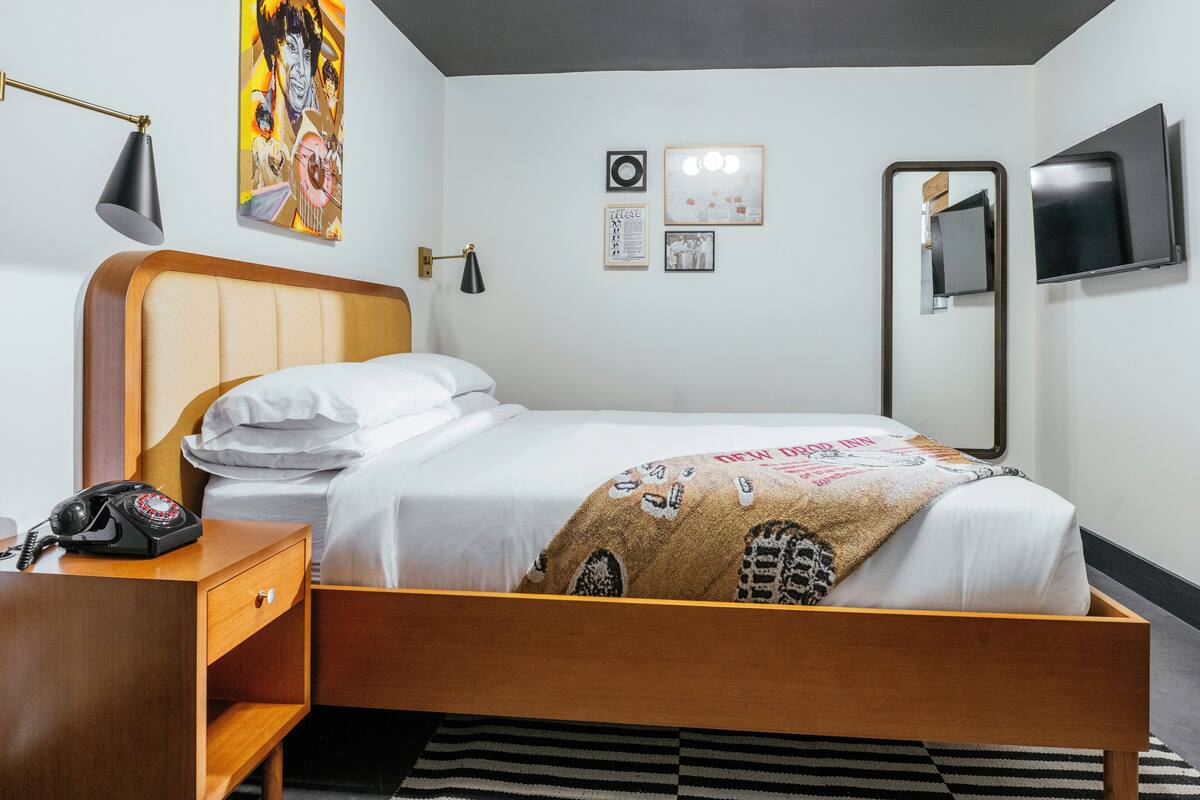 A hotel room with a wooden bed, a bedside telephone, a black lamp, and a wall-mounted TV.