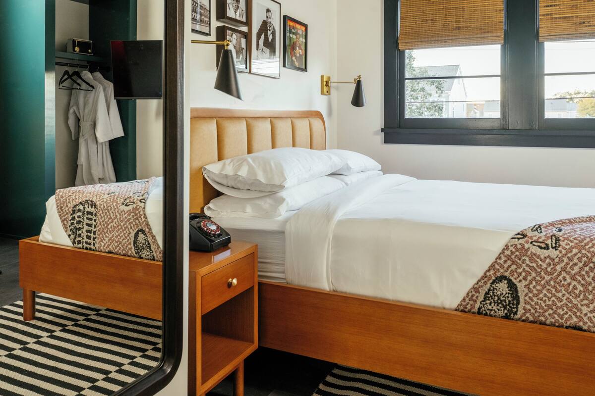 A bedroom with a wooden bed, a bedside table with a telephone, a mirror, and a window with blinds.
