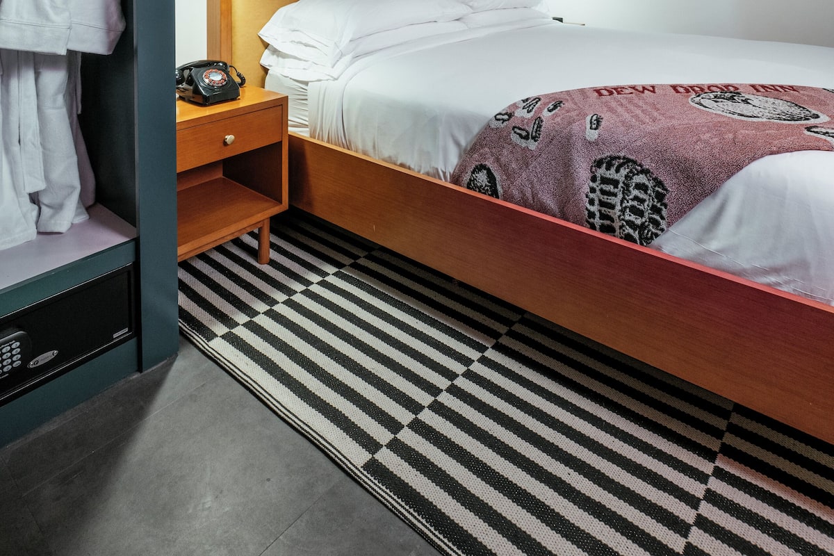 A hotel room with a bed, a nightstand, a wardrobe, a ceiling light fixture, and a black and white striped rug.