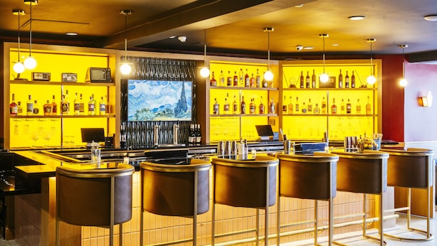 A bar area with multiple stools, a television screen, and shelves of bottles.