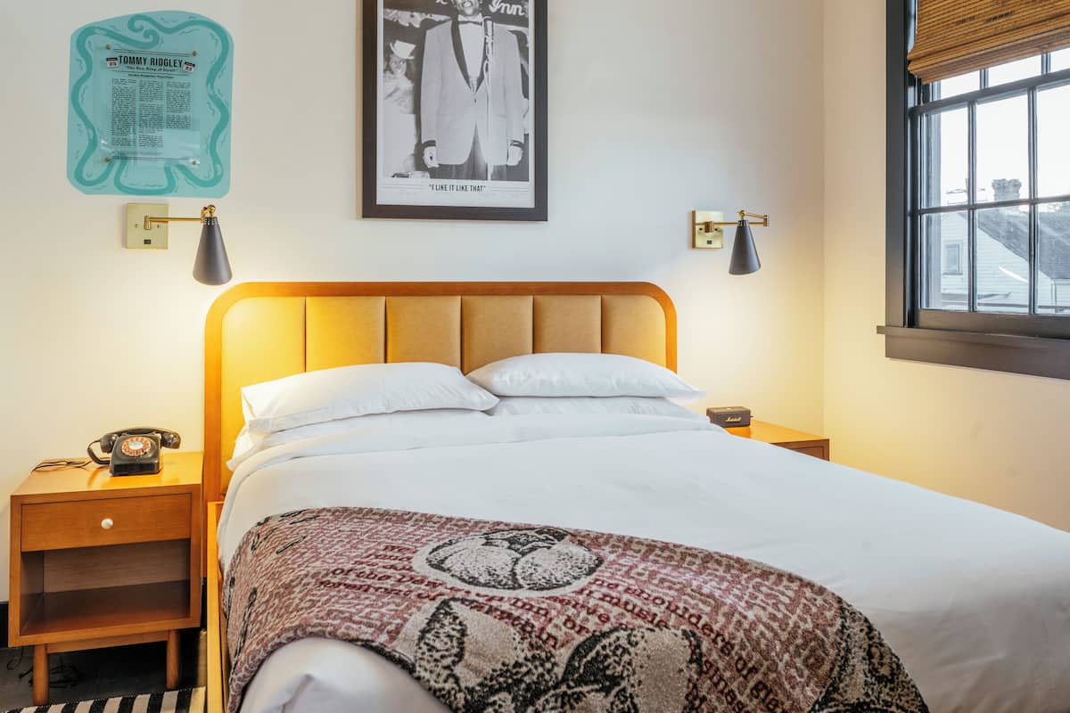 A neatly made bed with a patterned blanket, a wooden nightstand with a telephone, a framed picture on the wall, and a window with blinds.