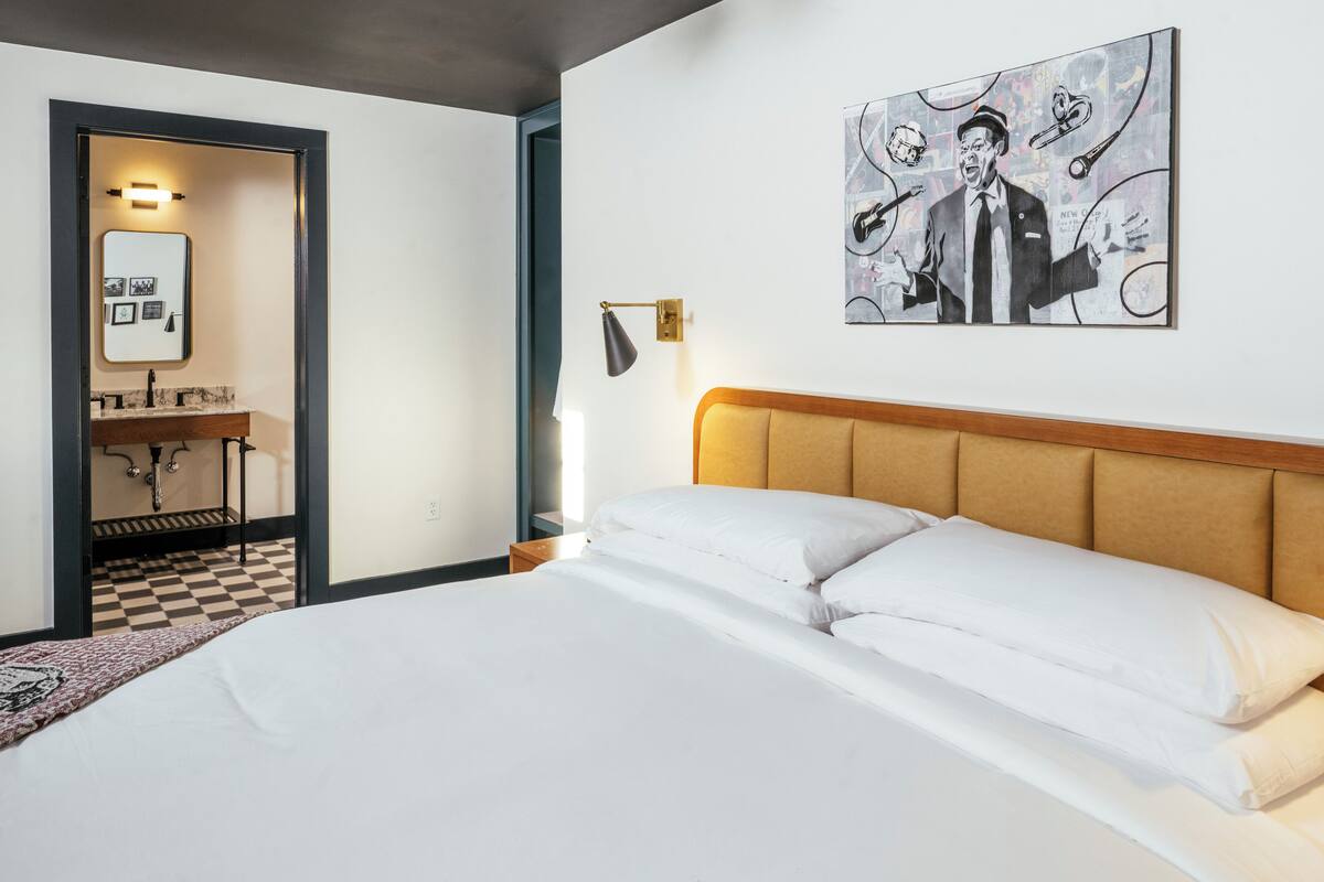 A hotel room with a large bed, a wooden headboard, a painting on the wall, and a desk with a mirror and lamp.