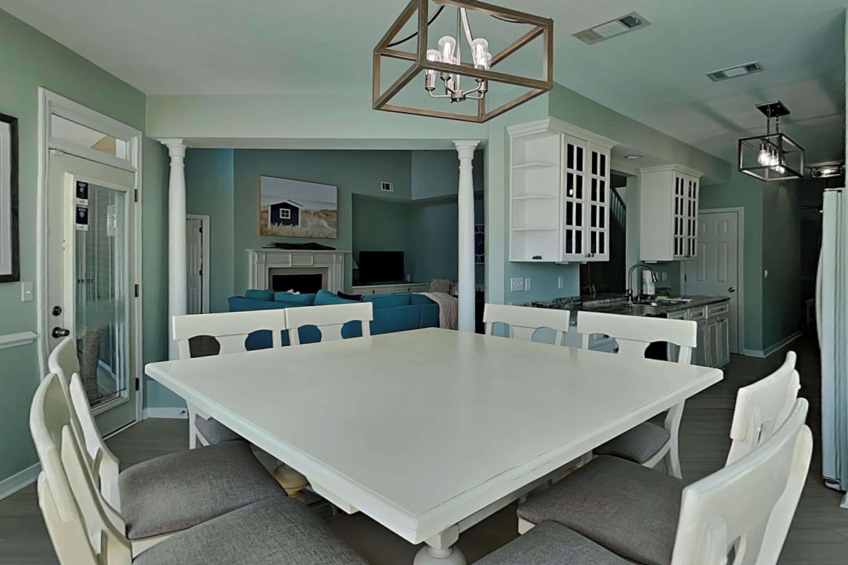 A dining room with a white table and chairs, a chandelier, and a kitchen in the background.