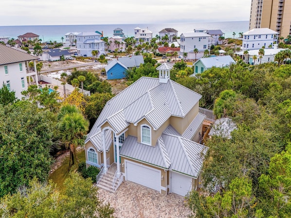 A coastal residential area with a prominent house featuring a unique roof design, surrounded by lush greenery and a view of the sea.