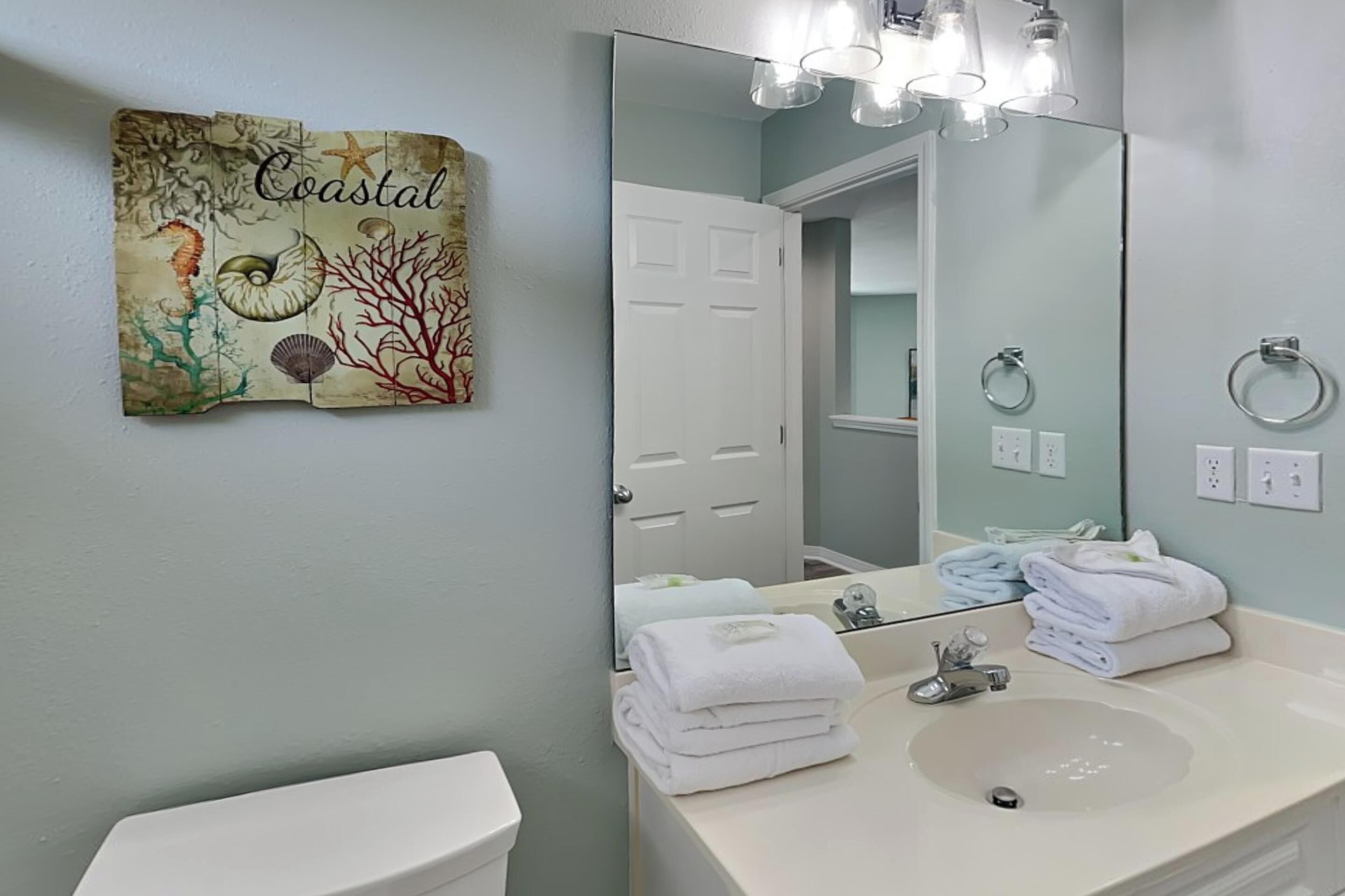 Un baño con un lavabo blanco, un espejo grande y una obra de arte en la pared titulada 'Coastal'.