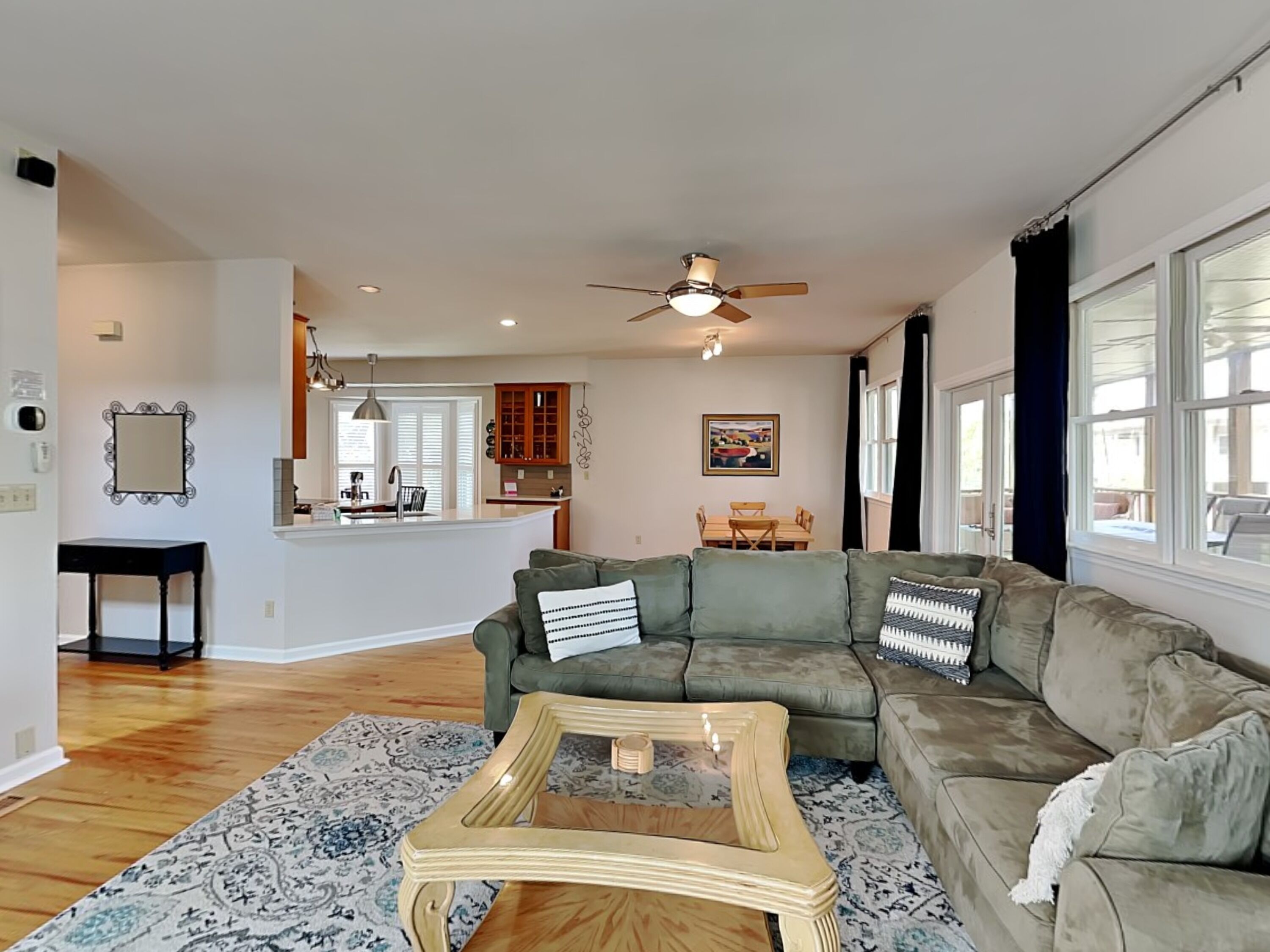 A spacious living room with a green sectional sofa, a coffee table, and a patterned rug. A kitchen with wooden cabinets and a dining area with a table and chairs are visible in the background.