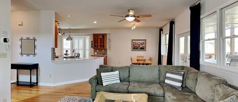 A spacious living room with a green sectional sofa, a coffee table, and a patterned rug. A kitchen with wooden cabinets and a dining area with a table and chairs are visible in the background.