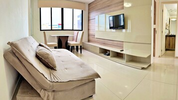 Uma sala de estar moderna com sofá, televisão na parede, área de jantar com cadeiras e vista através de uma janela ampla.