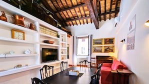 A furnished room with a dining table, chairs, a television, shelves with decorative items, a red sofa, and a fireplace. - Elegance in Rome's Heart - Smart Lodging Prime (Roma)