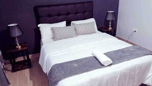 Ein ordentlich bezogenes Bett mit weißer Daunendecke, zwei gemusterten Kissen und einer grauen Tagesdecke. An jeder Seite des Bettes befinden sich Nachttische mit Lampen.