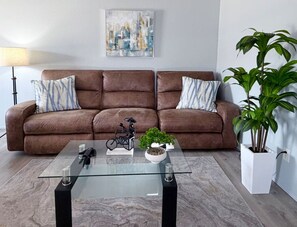 A brown leather sofa with striped pillows, a glass coffee table with decorative items, a potted plant, and a painting on the wall.