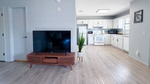 Sala de estar moderna com televisão de tela plana em um suporte de madeira, um vaso de planta e uma cozinha ao fundo.