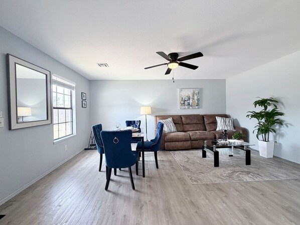 A living room with a dining area, a ceiling fan, a large window, and a potted plant.