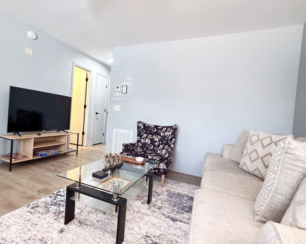A living room with a flat-screen TV, a glass coffee table, a floral armchair, and a beige sofa.