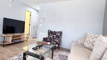 A living room with a flat-screen TV, a glass coffee table, a floral armchair, and a beige sofa.