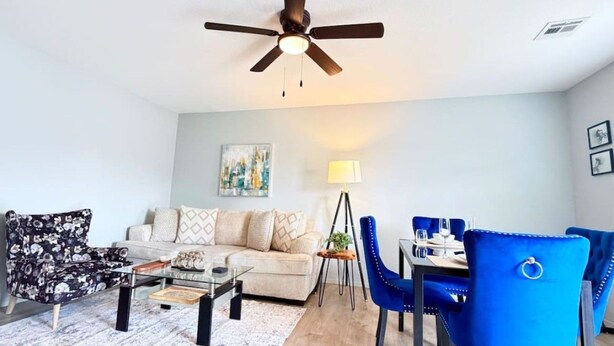 Een moderne woonkamer met een plafondventilator, een zithoek, een salontafel en een eethoek met blauwe stoelen.