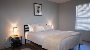 Een slaapkamer met een bed, nachtlampjes, een vloerkleed, een lijst met een foto aan de muur en een raam met zonwering.
