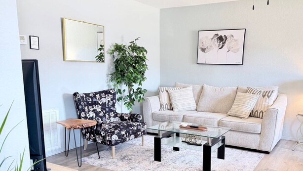 A living room with a sofa, a glass coffee table, a floral armchair, a potted plant, and a television. - Townhome, Walk to The Momentary and Trails (Bentonville)
