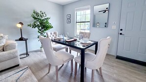 Uma sala de jantar com mesa de vidro, cadeiras estofadas brancas, planta em vaso e luminária moderna.